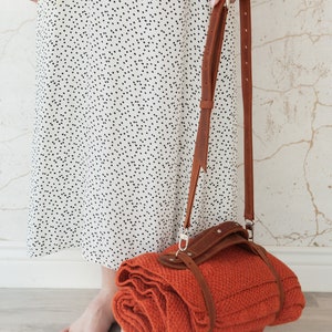 May include: A rolled orange blanket with a brown leather strap and shoulder strap. The blanket is held by a person wearing a white dress with black polka dots. The background is a light-colored wall.