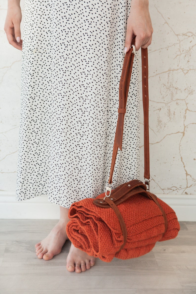 May include: A rolled orange blanket with a brown leather strap and shoulder strap. The blanket is held by a person wearing a white dress with black polka dots. The background is a light-colored wall.