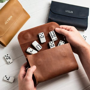 May include: A brown leather domino set with white dominoes featuring black dots. The set is in a brown leather case, with a tan leather case and a navy blue leather case in the background. The tan case has the name "ALYSON" embossed on it.