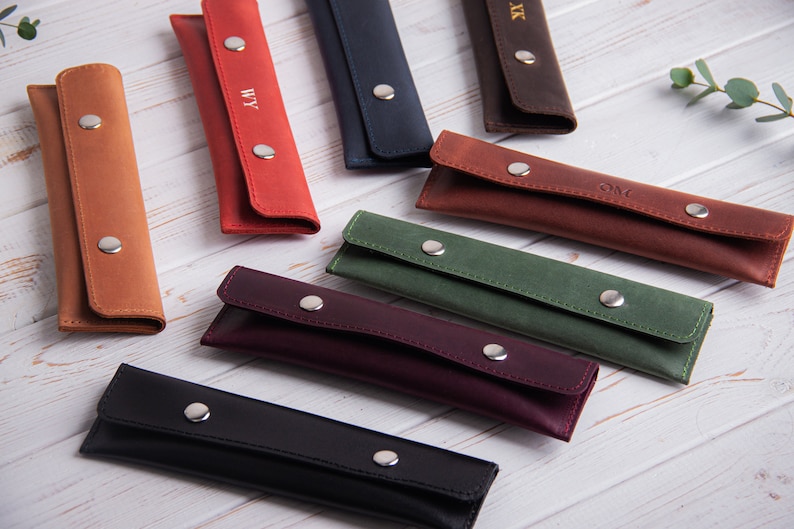 May include: Nine leather pencil cases in various colors, including brown, red, blue, green, purple, and black. Each case has a flap with a snap closure and is personalized with initials.