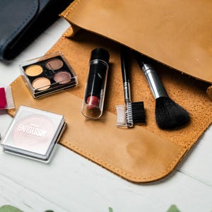 May include: A tan leather makeup bag open to reveal various cosmetic products. Items include an eyeshadow palette, lipstick, a blush compact, a makeup brush, and other applicators. A dark blue wallet is visible in the background.