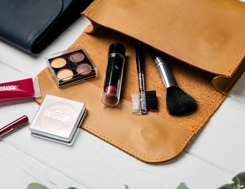 May include: A tan leather makeup bag open to reveal various cosmetic products. Items include an eyeshadow palette, lipstick, a blush compact, a makeup brush, and other applicators. A dark blue wallet is visible in the background.