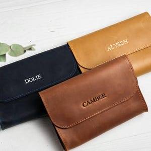 May include: Three leather wallets in navy blue, tan, and brown. Each wallet has a flap closure and is embossed with a name: "Dolie", "Alyson", and "Camber". The wallets are arranged on a white wooden surface.