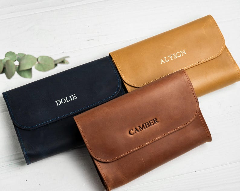 May include: Three leather wallets in navy blue, tan, and brown. Each wallet has a flap closure and is embossed with a name: "Dolie", "Alyson", and "Camber". The wallets are arranged on a white wooden surface.