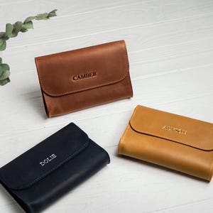 May include: Three leather wallets in shades of brown, navy blue, and tan are displayed on a white wooden surface. Each wallet has a flap closure and is embossed with a name: "CAMBER", "DOLIE", and "ALYSON".