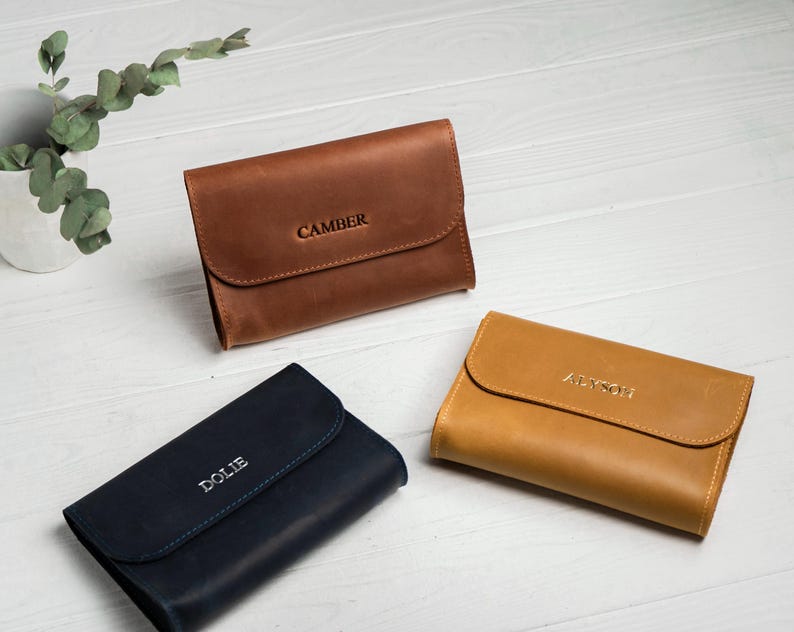 May include: Three leather wallets in shades of brown, navy blue, and tan are displayed on a white wooden surface. Each wallet has a flap closure and is embossed with a name: "CAMBER", "DOLIE", and "ALYSON".