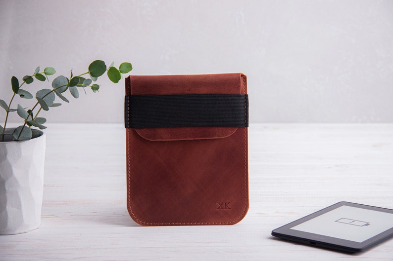 Puede incluir: Una funda de cuero marr&oacute;n con una banda el&aacute;stica negra y las iniciales "XK" grabadas en la parte delantera. La funda est&aacute; dise&ntilde;ada para contener un lector electr&oacute;nico.