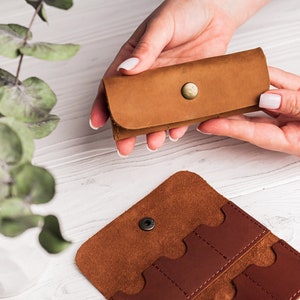 K&ouml;nnte beinhalten: Ein braunes Lederetui mit Druckknopfverschluss zur Aufbewahrung von Speicherkarten. Das Etui ist ge&ouml;ffnet und zeigt vier Steckpl&auml;tze f&uuml;r Speicherkarten. Eine einzelne Speicherkarte ist auf dem Tisch sichtbar.