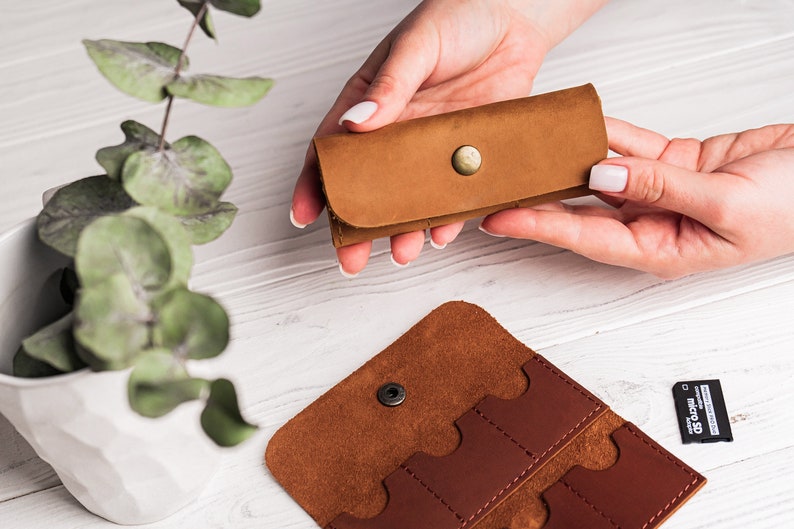 K&ouml;nnte beinhalten: Ein braunes Lederetui mit Druckknopfverschluss zur Aufbewahrung von Speicherkarten. Das Etui ist ge&ouml;ffnet und zeigt vier Steckpl&auml;tze f&uuml;r Speicherkarten. Eine einzelne Speicherkarte ist auf dem Tisch sichtbar.