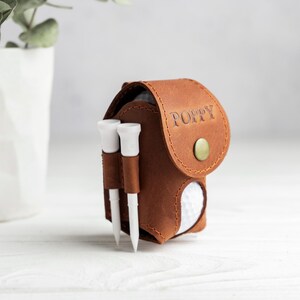 May include: A brown leather golf ball pouch with a flap closure secured by a brass-colored snap button. The name POPPY is embossed on the flap. Two white golf tees are held in loops on the side, and a white golf ball is visible inside.