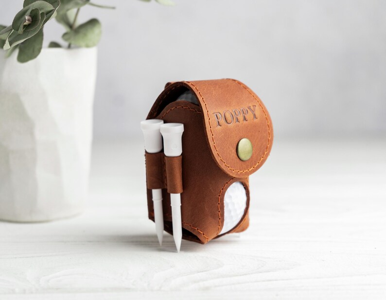 May include: A brown leather golf ball pouch with a flap closure secured by a brass-colored snap button. The name POPPY is embossed on the flap. Two white golf tees are held in loops on the side, and a white golf ball is visible inside.