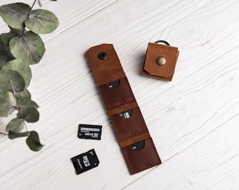 Könnte beinhalten: Ein braunes Lederetui mit drei Steckplätzen für Speicherkarten. Das Etui hat einen Schlüsselring. Zwei Speicherkarten sind sichtbar, eine mit der Aufschrift "micro SD Adapter" und die andere mit der Aufschrift "micro SD".