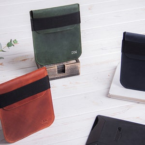 Puede incluir: Tres fundas de cuero para lectores electr&oacute;nicos en verde, marr&oacute;n y azul. Cada funda tiene una banda el&aacute;stica negra y est&aacute; personalizada con iniciales. Las fundas se muestran sobre una superficie de madera blanca.