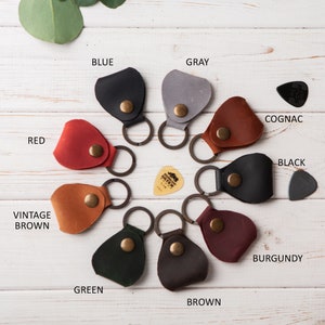 May include: Assortment of ten leather guitar pick holders in various colors, including red, blue, green, brown, gray, black, burgundy, cognac, and vintage brown. Each holder has a metal ring for attaching to a keychain.