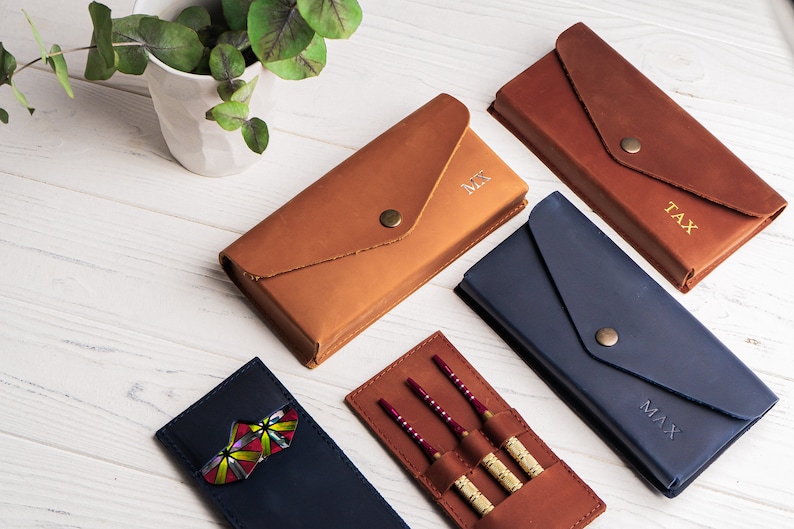 May include: Four leather cases for darts, each with a different personalized name: "Max", "Tax", "Mr", and a case with darts. The cases are brown and blue, with a flap closure.