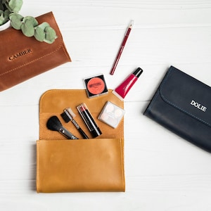 May include: An overhead shot of makeup and accessories on a white surface. There are three leather pouches, one brown with "CAMBER" embossed, one tan with makeup brushes and products, and one navy with "DOLIE" embossed. Makeup items include lipsticks, blush, and lip gloss.
