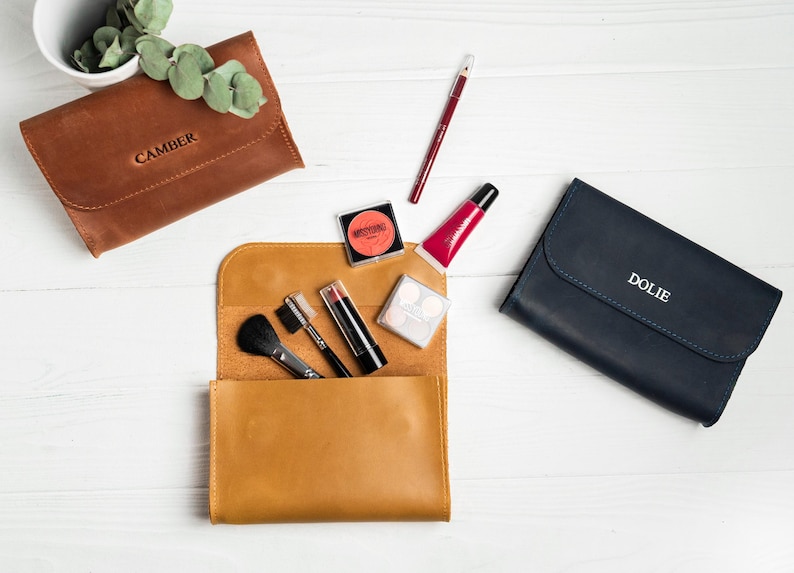 May include: An overhead shot of makeup and accessories on a white surface. There are three leather pouches, one brown with "CAMBER" embossed, one tan with makeup brushes and products, and one navy with "DOLIE" embossed. Makeup items include lipsticks, blush, and lip gloss.