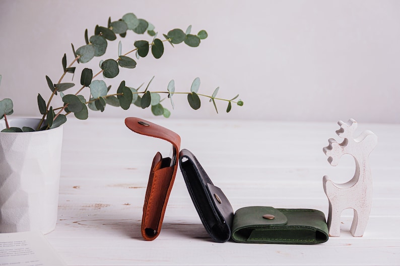 Pu&ograve; includere: Tre portafogli in pelle marrone, nero e verde, ciascuno con chiusura a bottone. Un cervo decorativo bianco e un vaso con rami di eucalipto sono anch'essi nell'immagine. I portafogli sono esposti su una superficie di legno bianco.