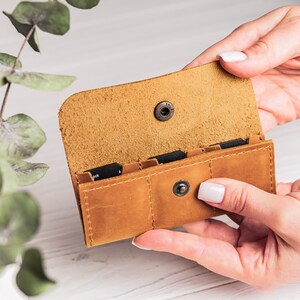 K&ouml;nnte beinhalten: Ein braunes Lederetui mit vier Steckpl&auml;tzen f&uuml;r Speicherkarten. Das Etui ist ge&ouml;ffnet und die Steckpl&auml;tze sind sichtbar. Das Etui wird in den H&auml;nden einer Person gehalten.