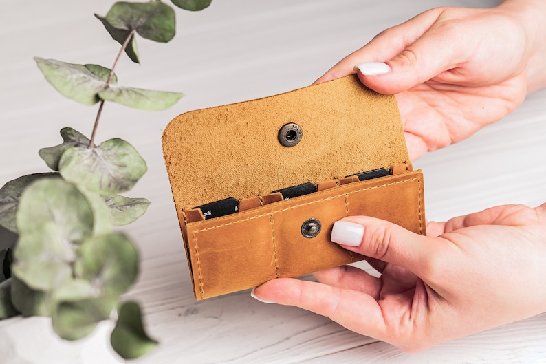 K&ouml;nnte beinhalten: Ein braunes Lederetui mit vier Steckpl&auml;tzen f&uuml;r Speicherkarten. Das Etui ist ge&ouml;ffnet und die Steckpl&auml;tze sind sichtbar. Das Etui wird in den H&auml;nden einer Person gehalten.