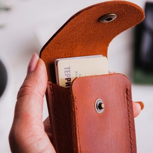 Pu&ograve; includere: Un porta carte in pelle marrone con chiusura a scatto, aperto per rivelare le carte all'interno. Il porta carte &egrave; tenuto in mano, mostrando il suo colore e la sua consistenza. Le carte hanno del testo. Un secondo porta carte nero &egrave; sullo sfondo.