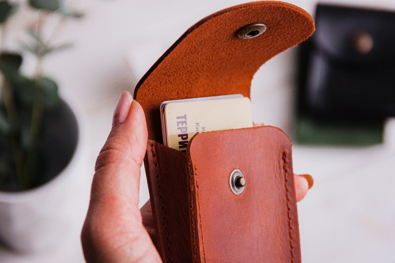 Pu&ograve; includere: Un porta carte in pelle marrone con chiusura a scatto, aperto per rivelare le carte all'interno. Il porta carte &egrave; tenuto in mano, mostrando il suo colore e la sua consistenza. Le carte hanno del testo. Un secondo porta carte nero &egrave; sullo sfondo.