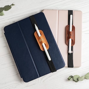 May include: A navy blue and a light pink tablet case, each featuring a black elastic band with a tan leather pen holder. The leather holders are embossed with the initials D.M. and B.N.B. respectively, and each holds a white stylus pen.