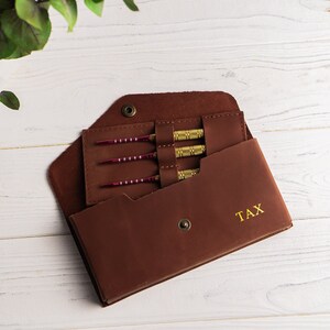 May include: A brown leather dart case with gold stitching and the word "TAX" embossed on the front. The case is open, revealing four darts with red and gold accents.