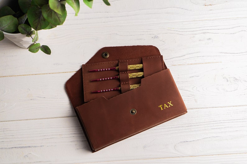 May include: A brown leather dart case with gold stitching and the word "TAX" embossed on the front. The case is open, revealing four darts with red and gold accents.
