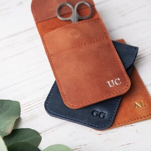 May include: Three leather pouches with initials 'UC', 'GG', and 'AL' embossed on them. The pouches are stacked on top of each other. The top pouch has a pair of small scissors inside.
