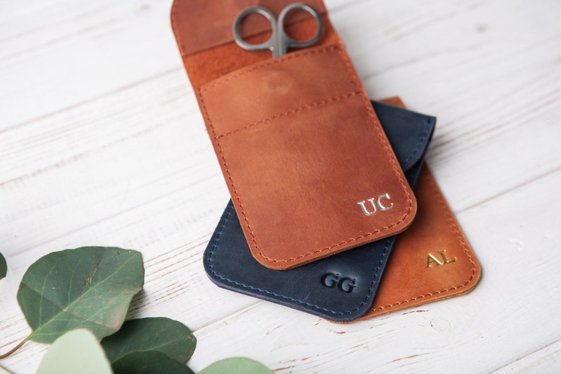 May include: Three leather pouches with initials 'UC', 'GG', and 'AL' embossed on them. The pouches are stacked on top of each other. The top pouch has a pair of small scissors inside.