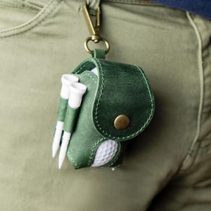 May include: A green leather golf accessory pouch with a brass clasp, holding a golf ball and two golf tees. The pouch is attached to a belt loop. The golf ball is white with dimples.