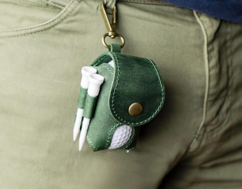 May include: A green leather golf accessory pouch with a brass clasp, holding a golf ball and two golf tees. The pouch is attached to a belt loop. The golf ball is white with dimples.