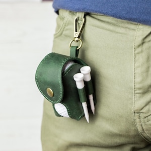 May include: A dark green leather golf ball pouch with a brass snap closure and a brass swivel clip. The pouch holds a white golf ball and features two exterior side loops, each containing a white golf tee. The pouch is attached to a belt loop.