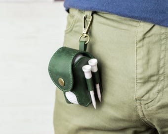 May include: A dark green leather golf ball holder with a gold-colored clasp, holding a white golf ball and two golf tees. The holder is attached to a belt loop. The golf tees are white with green tips.