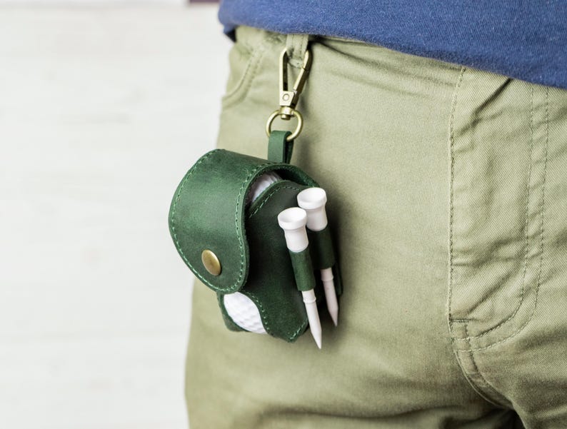 May include: A dark green leather golf ball pouch with a brass snap closure and a brass swivel clip. The pouch holds a white golf ball and features two exterior side loops, each containing a white golf tee. The pouch is attached to a belt loop.