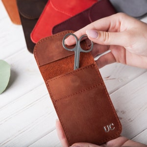 May include: A brown leather case with a small pocket for a pair of scissors. The case has a flap that closes with a snap and has the initials "UC" embossed on the front.