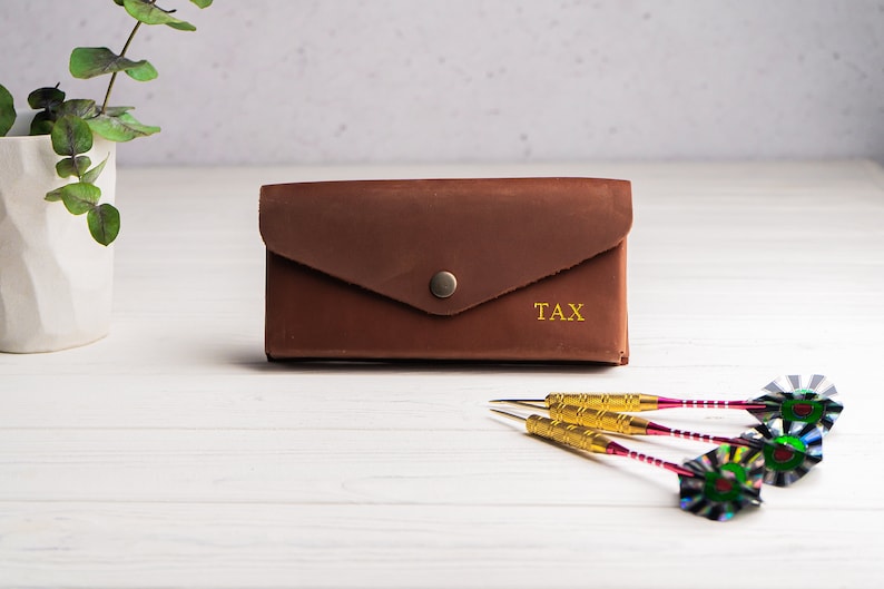 May include: A brown leather wallet with a flap and a gold button. The wallet has the word "TAX" embossed on the front. Three gold darts with colorful fletching are on a white wooden surface.