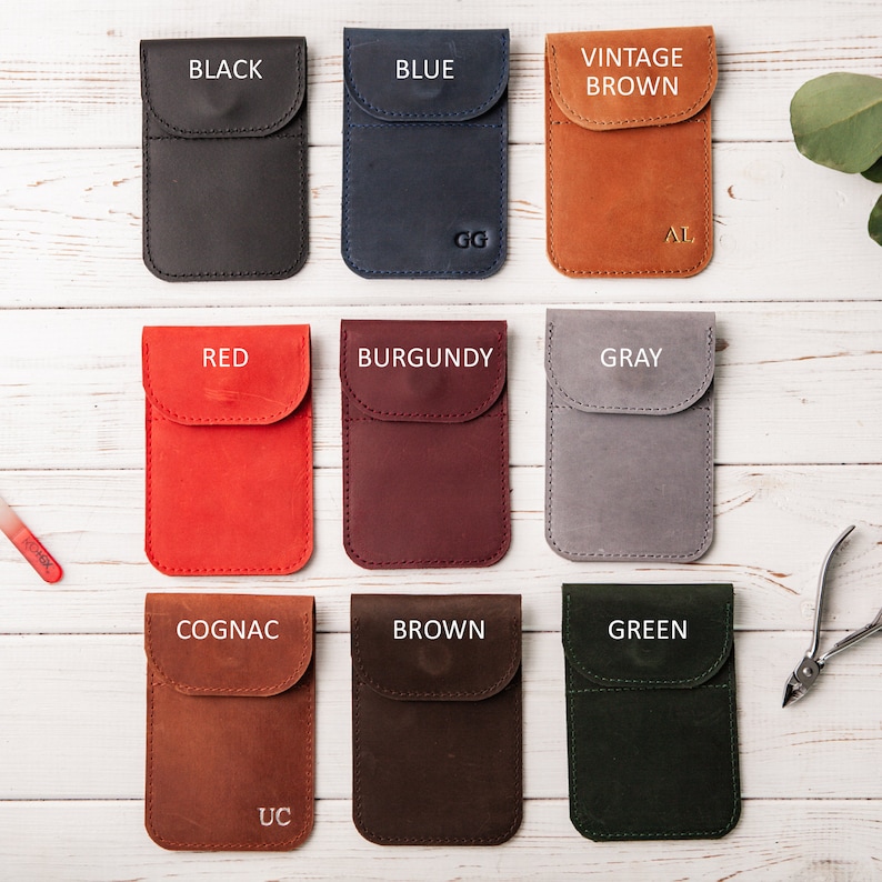 May include: Twelve leather phone cases in various colors, including black, blue, vintage brown, red, burgundy, gray, cognac, brown, and green. Each case has a flap with a stitched edge and a small pocket on the back. The cases are arranged in three rows of four on a white wooden surface.