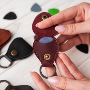May include: A burgundy leather guitar pick holder with a snap closure and a key ring. The holder is open and a blue guitar pick is visible inside.