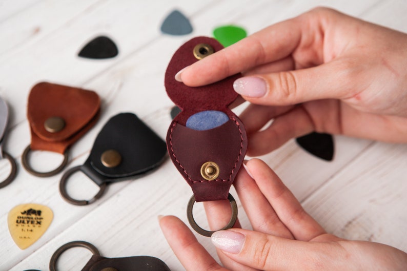May include: A burgundy leather guitar pick holder with a snap closure and a key ring. The holder is open and a blue guitar pick is visible inside.