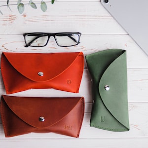 Könnte beinhalten: Eine Auswahl an Leder-Brillenetuis in Rot, Braun und Grün, jeweils mit Druckknopfverschluss. Das rote Etui hat die Initialen "SF", das braune "HM" und das grüne "EB". Eine schwarze Brille liegt darüber.