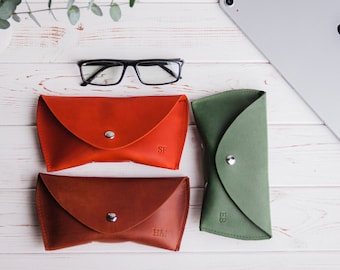 Könnte beinhalten: Eine Auswahl an Leder-Brillenetuis in Rot, Braun und Grün, jeweils mit Druckknopfverschluss. Das rote Etui hat die Initialen "SF", das braune "HM" und das grüne "EB". Eine schwarze Brille liegt darüber.