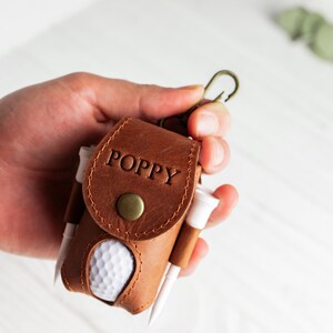 May include: A brown leather golf ball holder with the name "POPPY" embossed on the flap. The holder contains a golf ball and several golf tees. A brass-colored clasp is attached to the top.