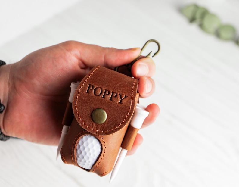 May include: A brown leather golf ball holder with the name "POPPY" embossed on the flap. The holder contains a golf ball and several golf tees. A brass-colored clasp is attached to the top.