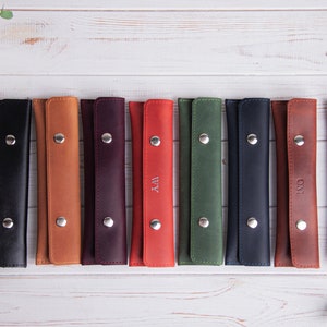 May include: Ten leather pencil cases in different colors, each with a silver button closure and personalized with initials. The cases are lined up on a white wooden surface.