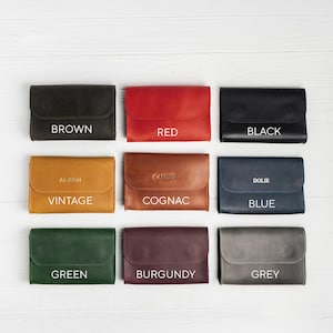 May include: Nine leather wallets in various colors, including brown, red, black, vintage, cognac, blue, green, burgundy, and grey. Each wallet has a flap closure and is labeled with its color name in white text.