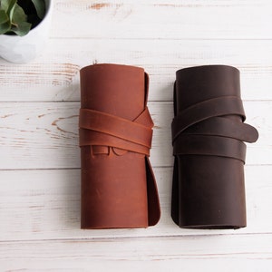 Op de afbeelding: Twee leren potloodkokers, een bruin en een donkerbruin, zijn opgerold en vastgebonden met een leren band. De kokers liggen op een wit houten oppervlak.