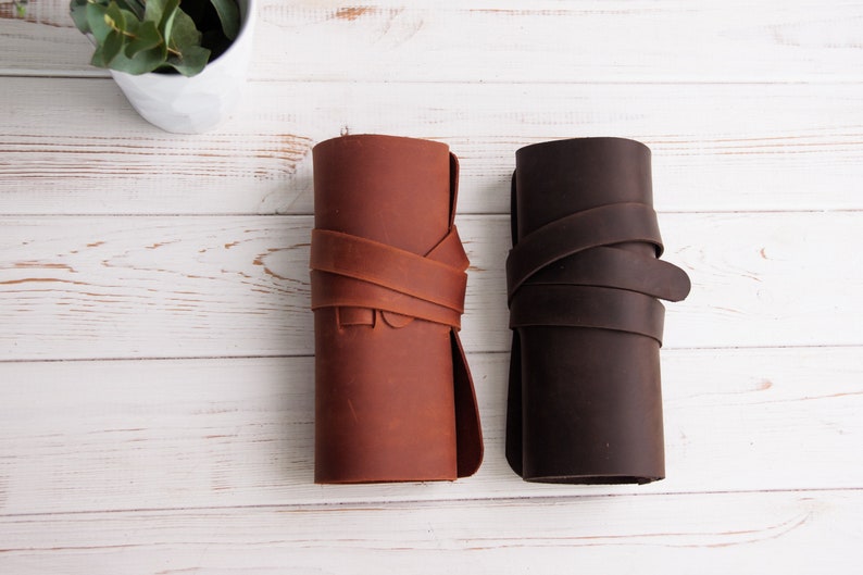Op de afbeelding: Twee leren potloodkokers, een bruin en een donkerbruin, zijn opgerold en vastgebonden met een leren band. De kokers liggen op een wit houten oppervlak.