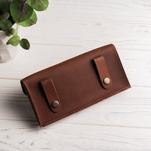 May include: A brown leather wallet with two snap closures. The wallet is lying on a white wooden surface.